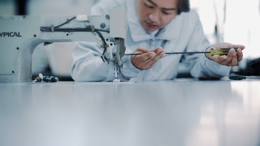 Un artigiano o tecnico si china su una macchina da cucire industriale, concentrato nella regolazione di un meccanismo con un cacciavite. L'immagine simboleggia la cura artigianale, la precisione della manifattura e l'applicazione della tecnologia avanzata nella creazione dei capi Cape Horn - Cape Horn